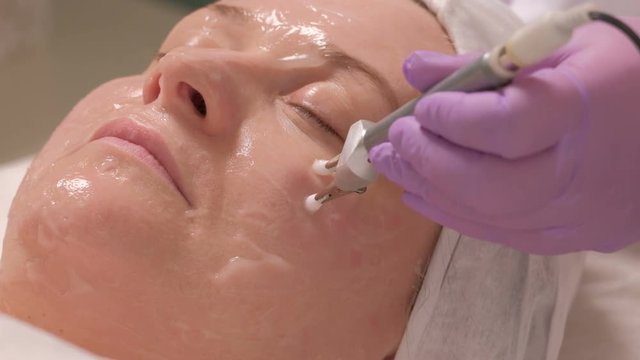 Woman receiving stimulating facial treatment with microcurrent equipment.Close-up plan of electrodes in the hands of a beautician, touching the female cheek through a mask. Wrinkle smoothing.