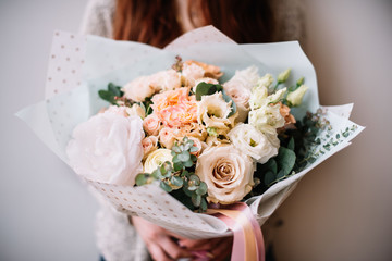 Very nice young woman holding blossoming flower bouquet of fresh peony, roses, eustoma, carnations in pastel cream and peach colors on the grey wall background