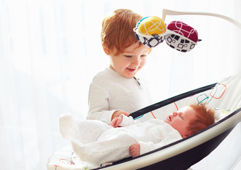 happy toddler baby brother welcoming his little baby sister, that lying in cradle swings