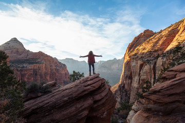 Adventurous Girl at the edge of a cliff is looking at a beautiful landscape view in the Canyon during a vibrant sunset. Taken in Zion National Park, Utah, United States.