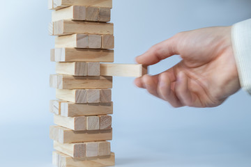 Man's hand taking the first block or putting the last block to a sturdy tower of wooden blocks. Concept photo of planning, taking risks and strategizing. Hand is in motion. Light gray background.