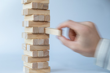 Man's hand taking the first block or putting the last block to a sturdy tower of wooden blocks. Concept photo of planning, taking risks and strategizing. Hand is in motion. Light gray background.