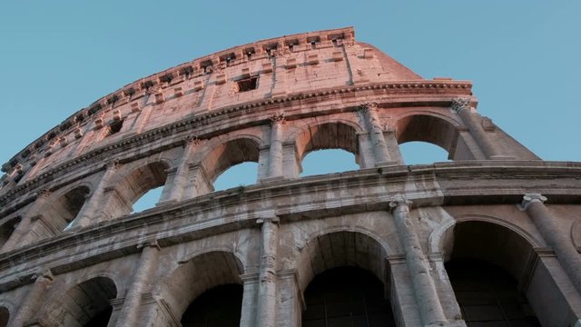 Camera tracking left low angle view of the Roman Colosseum facade with top floor arches and windows against a clear blue sky in Rome, Italy. 4K UHD at 29.97fps