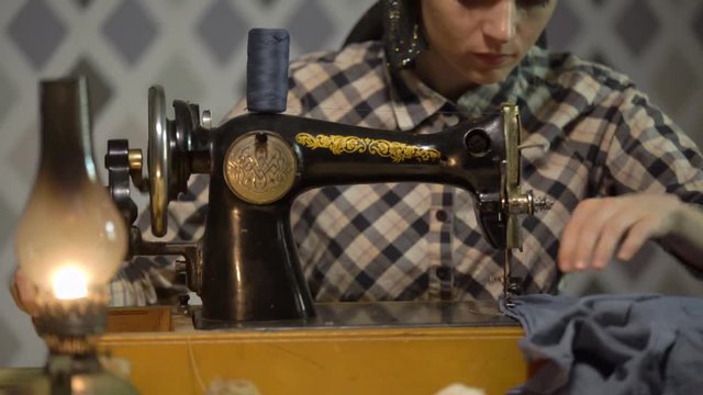 Retro seamstress girl sews cloth with old manual hand sewing machine. Woman works at home or workshop at night with kerosene lamp, listens music vinyl plate, gramophone or phonograph