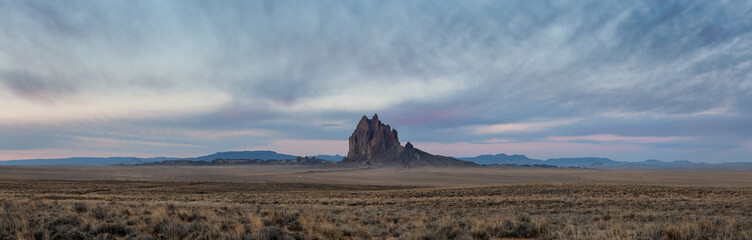 Dramatic panoramic landscape view of a dry desert with a mountain peak in the background during a vibrant cloudy sunrise.Taken at Shiprock, New Mexico, United States.