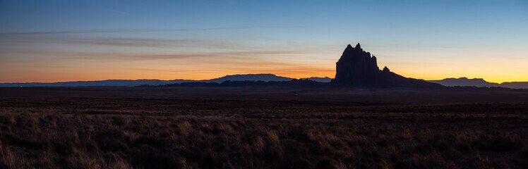 Dramatic panoramic landscape view of a dry desert with a mountain peak in the background during a vibrant sunset. Taken at Shiprock, New Mexico, United States.