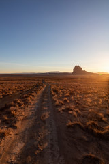 Striking landscape view of a dirt road in the dry desert with a mountain peak in the background during a vibrant sunset. Taken at Shiprock, New Mexico, United States.