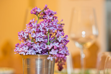Lilac flowers as spring bouquet in a metal cup on a table setting with wine glasses in the background. Warm shades in a cozy interior. Slight blur for dreamy effect.
