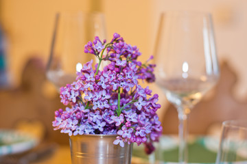 Lilac flowers as spring bouquet in a metal cup on a table setting with wine glasses in the background. Warm shades in a cozy interior. Slight blur for dreamy effect.