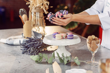 Close up female baker's hands with black manicure holding colorful french macaroons over a marble table with a plate, lavender and eucalyptus.
