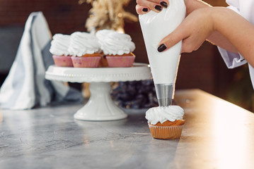 Close up hands of the female chef with confectionery bag squeezing cream on cupcakes.