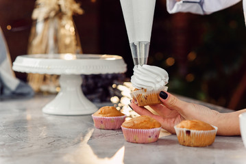 Close up hands of the female chef with confectionery bag squeezing cream on cupcakes.