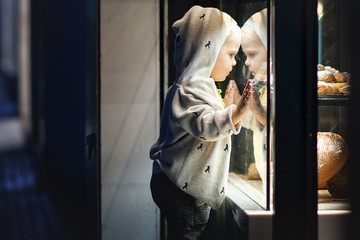 A little boy near the window of the bakery looking at the bread.