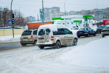 The car goes on a cleared road after heavy snowfall and snowstorms in the city. Traffic lights hang on the wire above the road. The included headlights at the car in the winter.