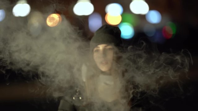 A girl is smoking vape outside in the evening. Nice colorful bokeh in front of the evening city.