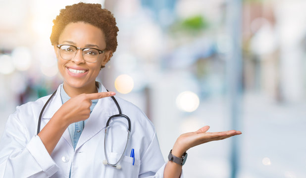 Young african american doctor woman wearing medical coat over isolated background amazed and smiling to the camera while presenting with hand and pointing with finger.