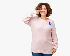 Middle ager senior woman wearing changeable blue color ribbon awareness over isolated background Pointing with hand finger to face and nose, smiling cheerful