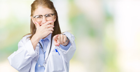 Middle age mature doctor woman wearing medical coat over isolated background Laughing of you, pointing to the camera with finger hand over mouth, shame expression