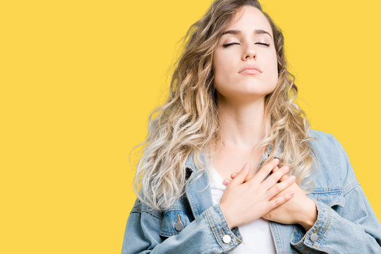 Beautiful young blonde woman wearing denim jacket over isolated background smiling with hands on chest with closed eyes and grateful gesture on face. Health concept.