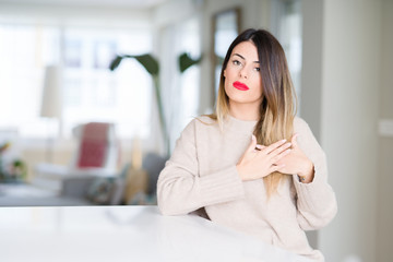 Young beautiful woman wearing winter sweater at home smiling with hands on chest with closed eyes and grateful gesture on face. Health concept.