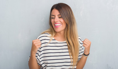 Obraz premium Young adult woman over grunge grey wall wearing navy t-shirt very happy and excited doing winner gesture with arms raised, smiling and screaming for success. Celebration concept.