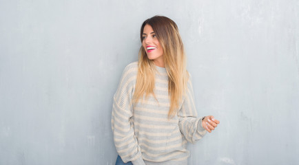Fototapeta premium Young adult woman over grunge grey wall wearing winter sweater looking away to side with smile on face, natural expression. Laughing confident.