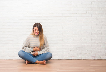 Young adult woman sitting on the floor over white brick wall at home with hand on stomach because indigestion, painful illness feeling unwell. Ache concept.