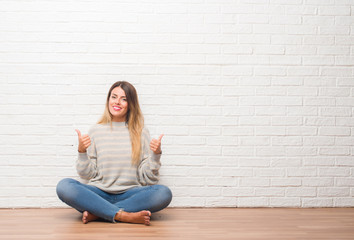 Young adult woman sitting on the floor over white brick wall at home success sign doing positive...