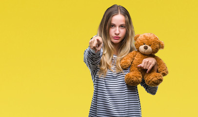 Young beautiful blonde woman holding teddy bear plush over isolated background pointing with finger to the camera and to you, hand sign, positive and confident gesture from the front