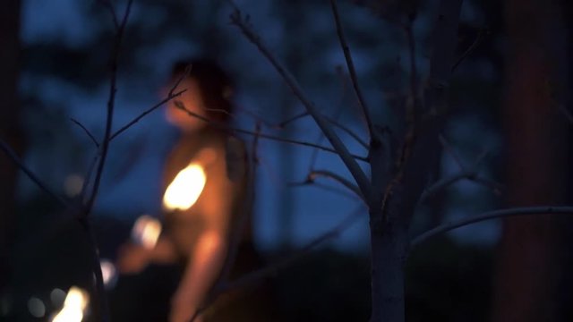 Slowmotion shot through trees of woman spinning fire fans at dusk