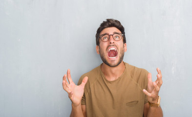 Handsome young man over grey grunge wall wearing glasses crazy and mad shouting and yelling with aggressive expression and arms raised. Frustration concept.