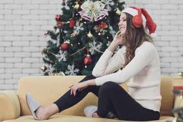 Young beautiful woman in red hat sitting on sofa between christmas trees and listening to music...