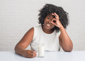 Young african american woman sitting on the table drinking a glass of milk with happy face smiling doing ok sign with hand on eye looking through fingers