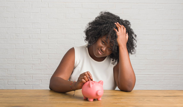 Young african american woman sitting on the table holding piggy bank stressed with hand on head, shocked with shame and surprise face, angry and frustrated. Fear and upset for mistake.