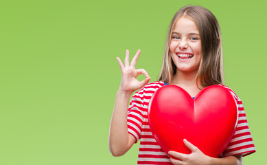 Young beautiful girl holding red heart over isolated background doing ok sign with fingers, excellent symbol