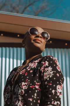 Bald African American Woman Wearing A Flower Patterned Bomber Jacket And Big Round Sunglasses.