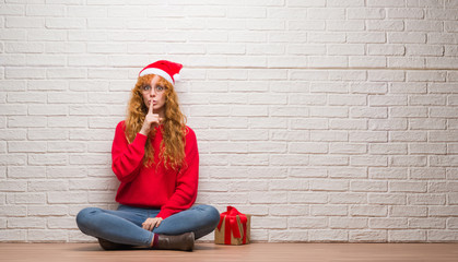 Young redhead woman sitting over brick wall wearing christmas hat asking to be quiet with finger on lips. Silence and secret concept.