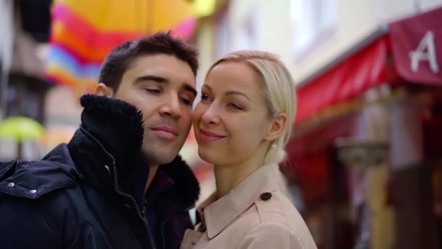 blonde woman is hugging with her husband on small city street, he is kissing her cheek