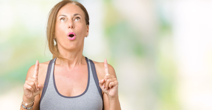 Beautiful middle age woman wearing sport clothes over isolated background amazed and surprised looking up and pointing with fingers and raised arms.