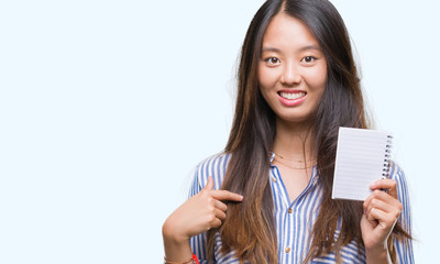 Young asian woman holding notebook over isolated background with surprise face pointing finger to himself