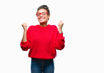 Fototapeta premium Young braided hair african american girl wearing sweater and glasses over isolated background celebrating surprised and amazed for success with arms raised and open eyes. Winner concept.