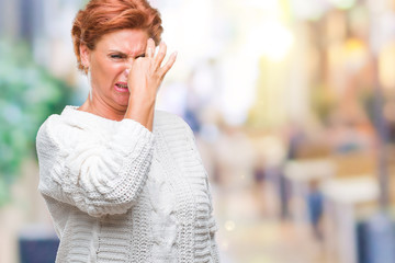 Atrractive senior caucasian redhead woman wearing winter sweater over isolated background smelling something stinky and disgusting, intolerable smell, holding breath with fingers on nose