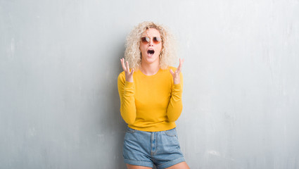 Fototapeta premium Young blonde woman with curly hair over grunge grey background crazy and mad shouting and yelling with aggressive expression and arms raised. Frustration concept.