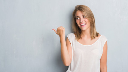 Beautiful young woman standing over grunge grey wall pointing and showing with thumb up to the side with happy face smiling