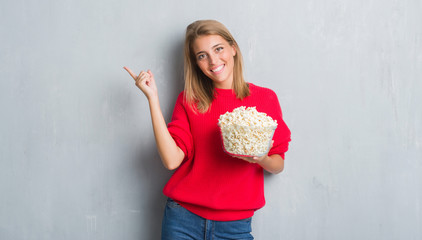 Beautiful young woman over grunge grey wall eating pop corn very happy pointing with hand and finger to the side