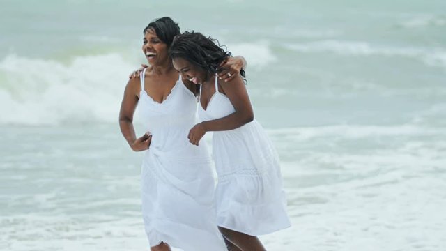 Happy African American mum and young daughter in white sun dress walking on beach vacation RED EPIC
