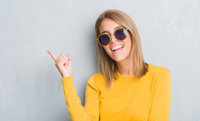 Beautiful young woman standing over grunge grey wall wearing retro sunglasses very happy pointing with hand and finger to the side