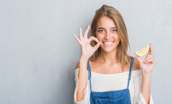 Beautiful young woman over grunge grey wall holding a lemon slice doing ok sign with fingers, excellent symbol