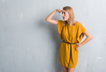 Fototapeta premium Beautiful young woman standing over grunge grey wall wearing a dress very happy and smiling looking far away with hand over head. Searching concept.