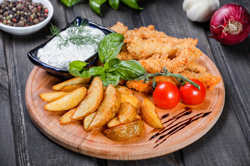 Breaded chicken, nuggets with roasted potato, sauce, tomatoes and greens on cutting board on dark wooden background